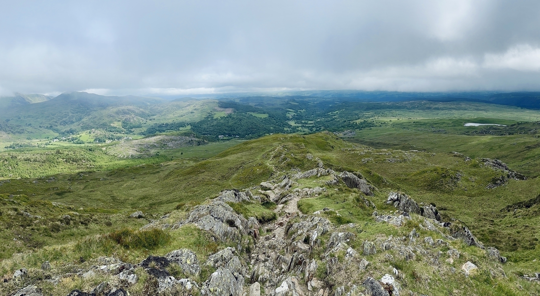 Hiking path down a hill, clouds cover the top third of the image and the valleys below and some other hills can be seen in the distance