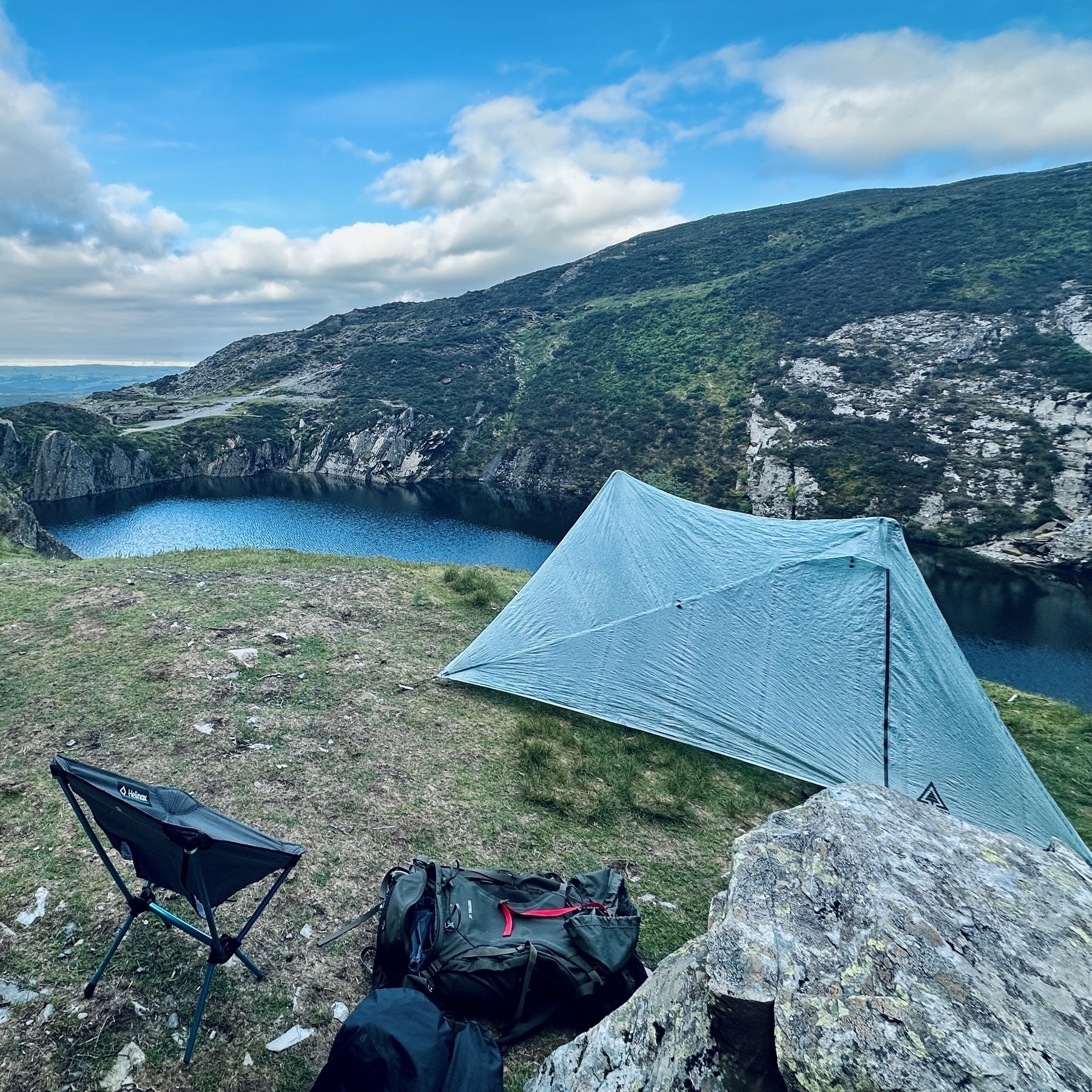 a trekking pole tent and a camping chair on grass near an unused quarry that has filled with water