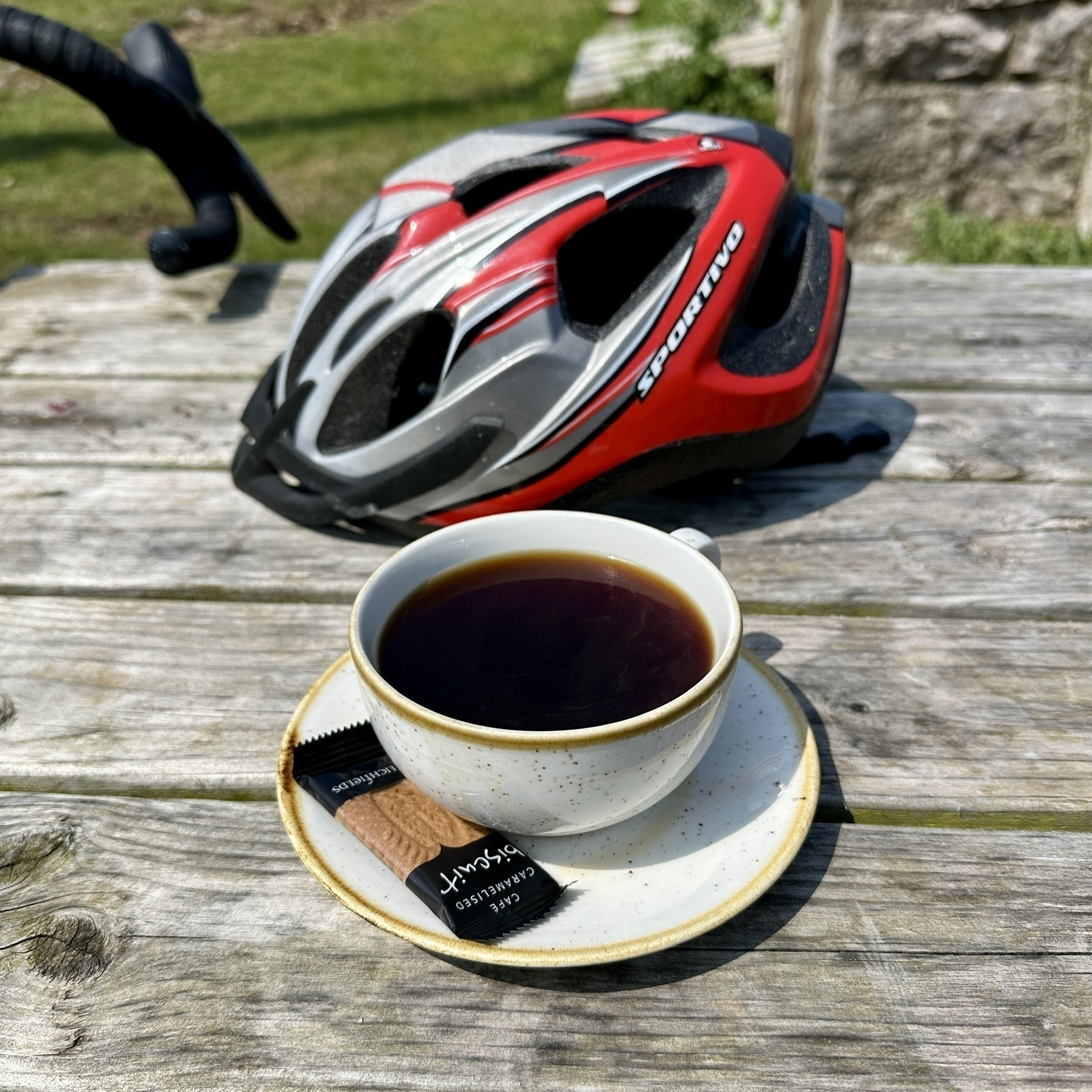 a black Americano coffee and a biscuit, along with a bike helmet, on a wooden bench at the Cafe at the top of the hill