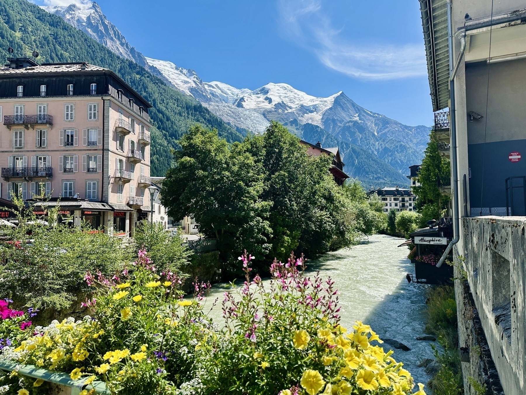 A scenic view of a mountain town features colorful flowers in the foreground, a river flanked by buildings, lush greenery, and snow-capped peaks under a clear blue sky.
