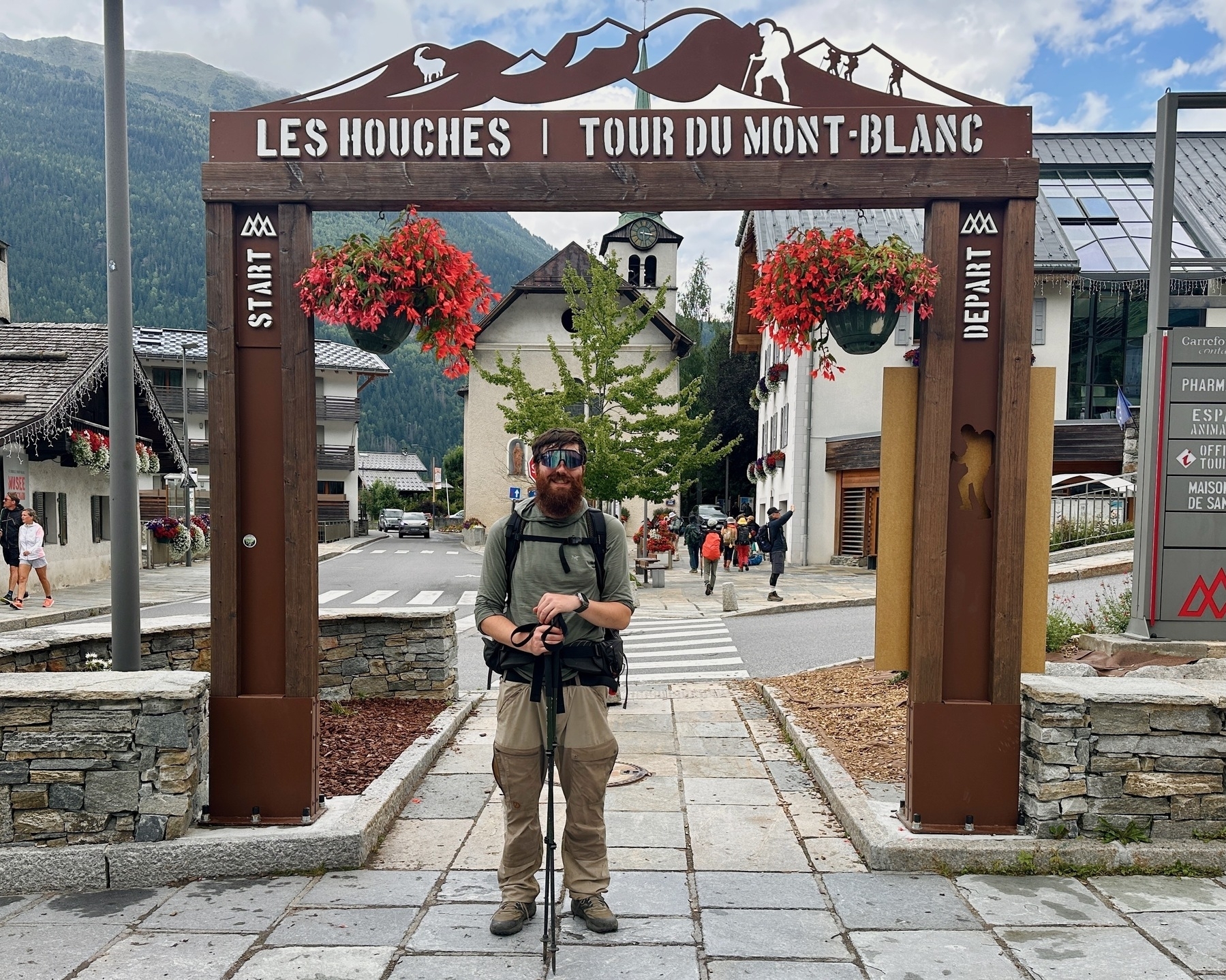 Me posing under the Les Houches Tour du Mont-Blanc archway, surrounded by mountainous scenery and buildings.