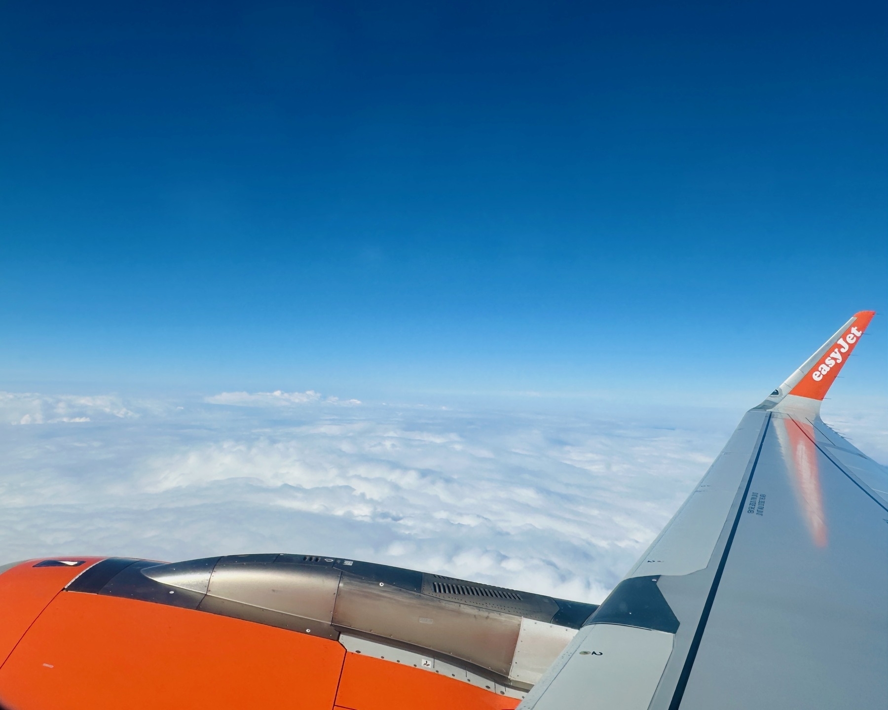 Auto-generated description: An airplane wing with orange accents is visible against a backdrop of blue sky and white clouds.